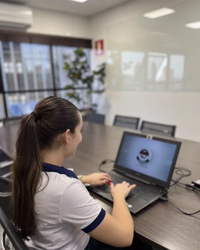 Colaboradora de costas atendendo no notebook, na sala de reuniões do escritório.