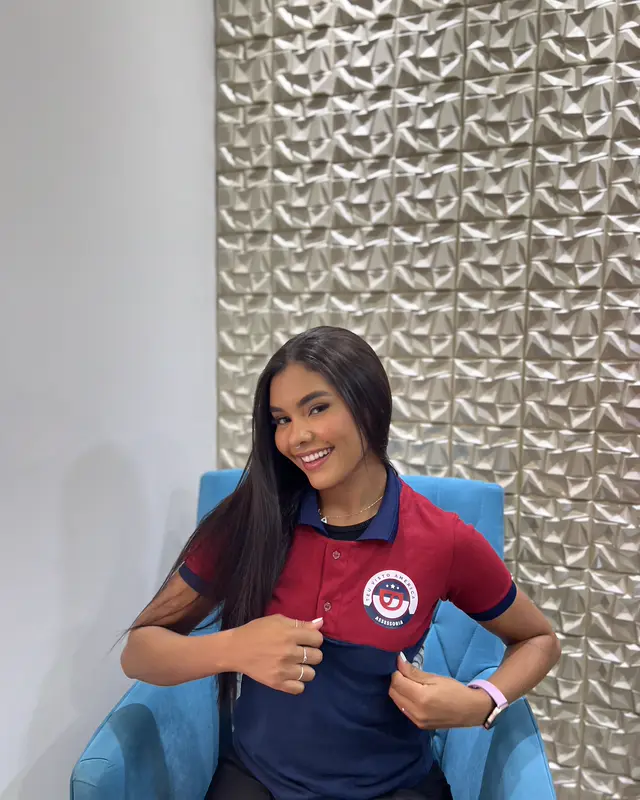 Colaboradora de uniforme sorrindo, sentada numa poltrona azul no escritório.