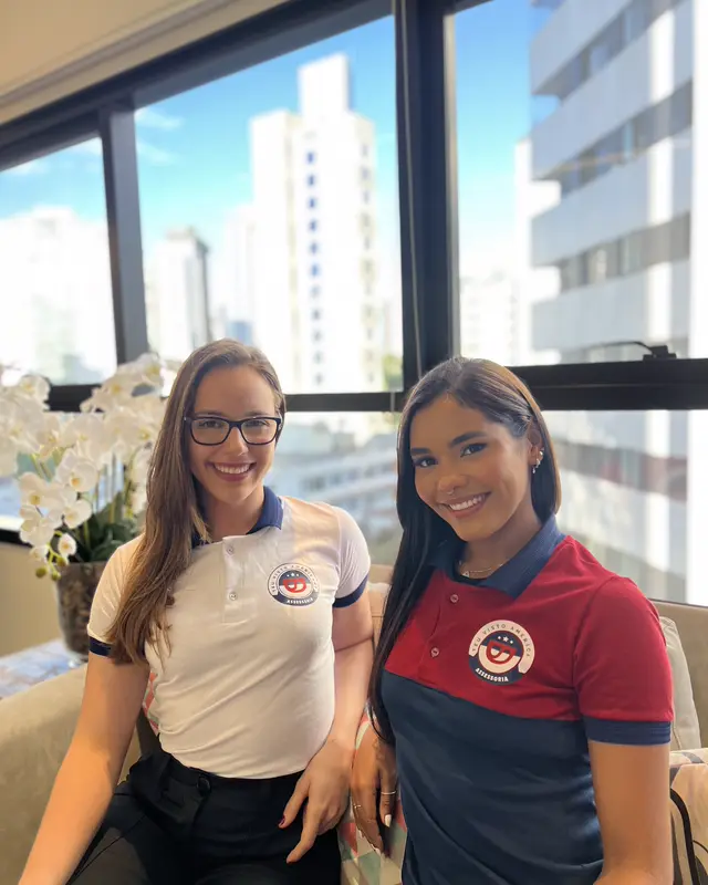Duas colaboradoras da Teu Visto Americano, de uniforme, no escritório com vista para a cidade.