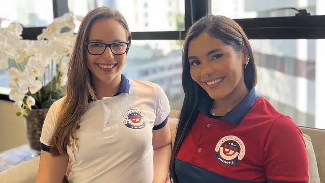 Duas colaboradoras da Teu Visto Americano, de uniforme, no escritório com vista para a cidade.